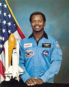 A formal NASA portrait of Ronald E. McNair, he proudly stands in front of a model of the shuttle, Challenger